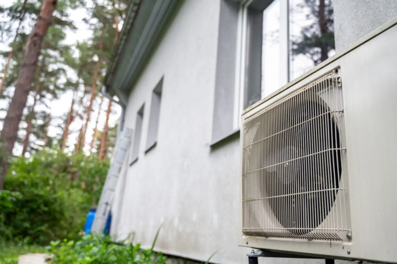 Udedel til varmepumpe på en hvid husmur med fyrretræer i baggrunden